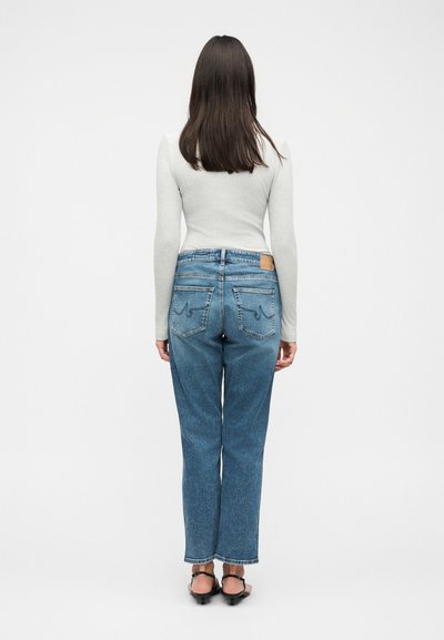 Γυναίκα με μακριά καστανά μαλλιά στέκεται με την πλάτη, φοράει ανοιχτό γκρι μπλούζα με μακριά μανίκια, μπλε τζιν και μαύρα ανοιχτά τακούνια σε λευκό φόντο.