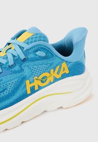 Baskets bleu ciel avec des accents jaunes, tige en mesh et une épaisse semelle blanche. Présente un tissu texturé et le logo HOKA bien en évidence.