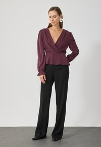 Blusa envolvente en color arándano con mangas largas y un dobladillo acampanado, combinada con pantalones negros ajustados. Tejido suave con un brillo sutil.