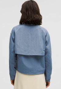 Vaaleansininen lyhyt takki, jossa on kerroksellinen selkäosa, valmistettu teksturoidusta kankaasta. Ominaisuuksiin kuuluu pitkät hihat ja elastiset hihansuut.