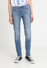 Jeans de denim azul de corte ajustado con un acabado desgastado, diseño de cinco bolsillos y pasadores para cinturón, combinados con zapatillas de lona negras.