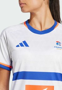 Valkoinen urheilupaita, jossa on sinisiä ja oransseja yksityiskohtia. Paita sisältää sinisen V-kaula-aukon, vaakasuoria raitoja ja Adidas-logon vasemmassa rinnassa.