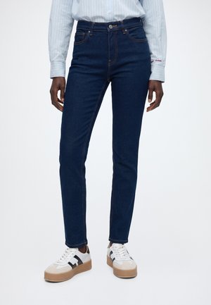 Jean slim taille haute bleu foncé porté avec des baskets blanches et beiges et une chemise rayée bleu clair sur une personne debout.