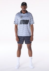 Grijs korte mouw shirt met kraag, voorzien van een geometrisch patroon en gedurfde zwarte letters. Gecombineerd met bijpassende donkere shorts en een zwarte pet.