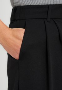 Une main repose à l'intérieur de la poche avant d'un pantalon noir plissé, assorti d'un haut gris côtelé.