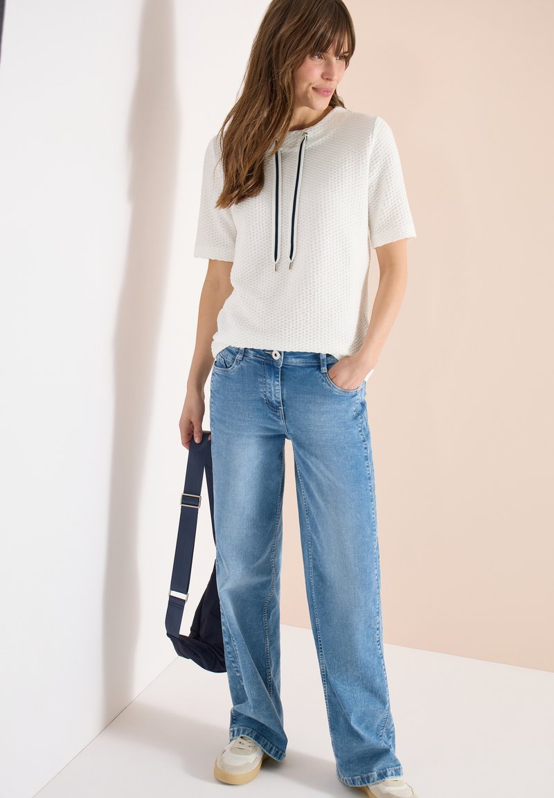 Femme portant un haut texturé blanc à manches courtes avec des cordons noirs, un jean large bleu clair, des baskets blanches, tenant un sac bleu marine à côté.