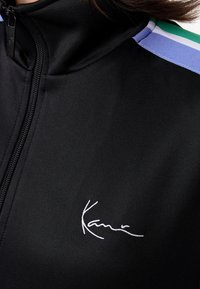 Juodas sportinis švarkas, pagamintas iš lygios medžiagos, pasižymintis aukštu apykakle, užtrauktuku ir balta išsiuvinėta logotipu priekyje. Akcentinės juostelės žalios ir mėlynos spalvos.