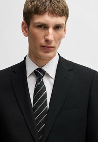 Abito nero con finitura testurizzata, camicia bianca e cravatta nera a righe con accenti argentati. Vista ravvicinata della parte superiore del corpo.