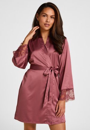 Femme aux longs cheveux foncés portant un peignoir en satin rose avec des poignets en dentelle noué à la taille sur un fond clair uni.