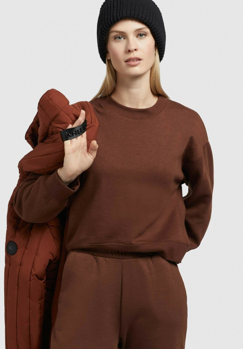 Eine Frau trägt eine schwarze Strickmütze sowie einen braunen Pullover und Hosen und hält eine rostfarbene gesteppte Jacke über ihrer Schulter.