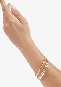 Tre bangles metalliche su un polso: oro, oro rosa e argento, con finiture texture e lisce, che riflettono la luce su uno sfondo neutro.
