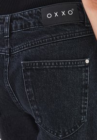 Jeans de mezclilla negra con corte recto, que cuentan con un bolsillo trasero visible, etiqueta de logo OXXO y un sutil desgaste ligero en la tela.