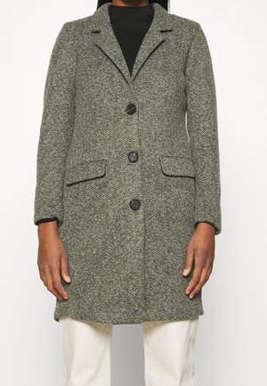 Kobieta ubrana w szary, teksturowany płaszcz do kolan z czarnymi guzikami, na czarnej bluzce i białych spodniach, ręce luźno opuszczone wzdłuż ciała.