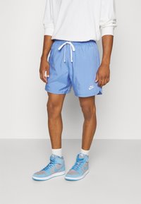 Muž na sobě má světle modré Nike šortky s bílou šňůrkou, bílé tričko s dlouhým rukávem, bílé ponožky a modré vysoké tenisky Nike.