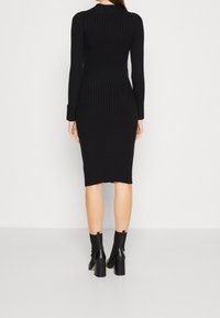Donna che indossa un abito midi nero a coste, aderente e con maniche lunghe, e stivali neri con tacco alla caviglia, in piedi su uno sfondo bianco semplice.