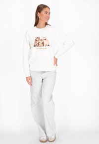 Fehér pulóver elöl három macskával és az "I'm holding tight" felirattal. Világos farmerrel és bézs cipővel párosítva.