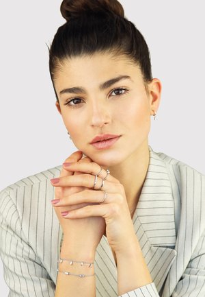 Silver jewelry featuring rings and a bracelet with geometric accents, worn on a model with a striped blazer and neutral background.