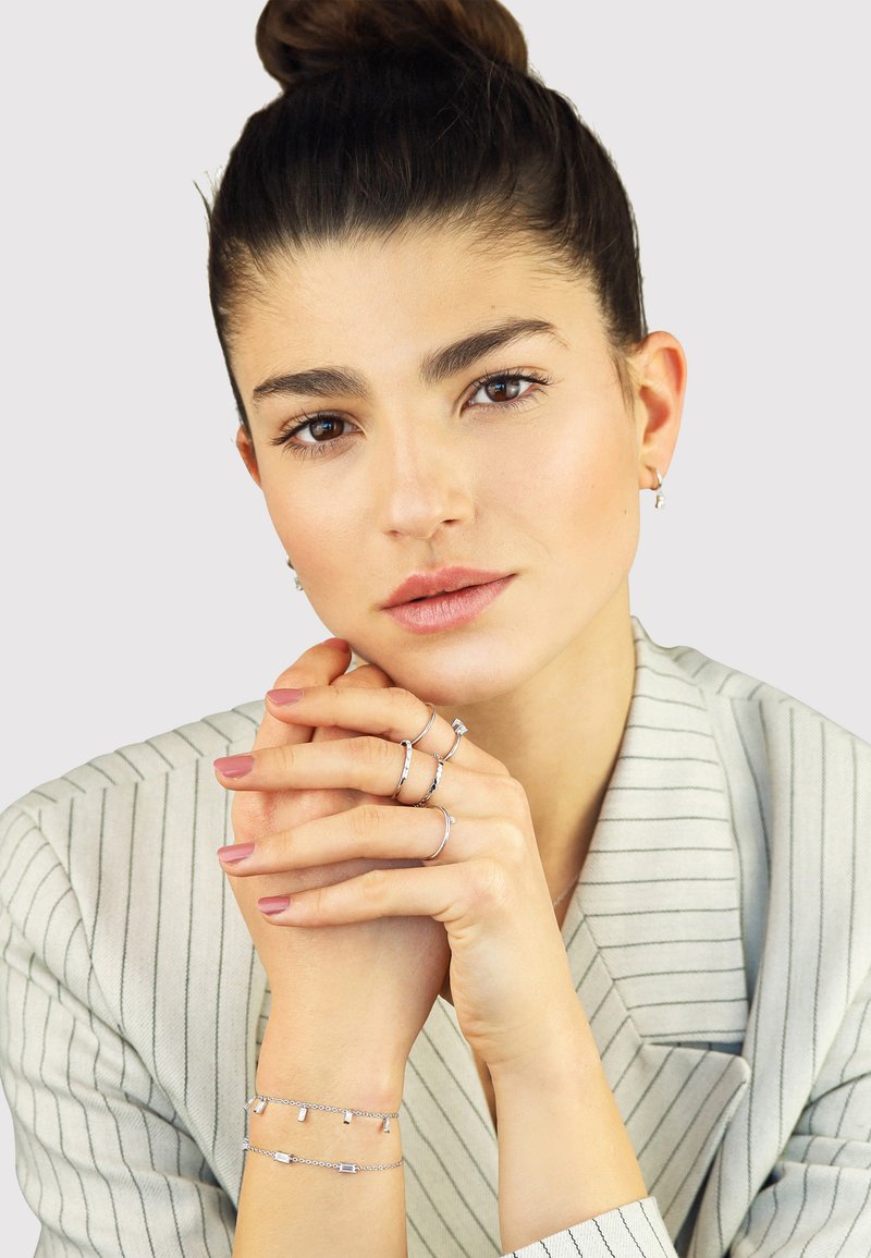 Silver jewelry featuring rings and a bracelet with geometric accents, worn on a model with a striped blazer and neutral background.