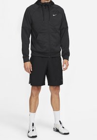 Sudadera negra con cremallera y cordones, bolsillos delanteros y logotipo de Nike. Combinada con pantalones cortos negros, calcetines blancos y zapatillas deportivas negras.