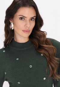 Groene gebreide trui met een opstaande kraag, versierd met verschillende gekleurde edelsteenachtige accenten. Lange, golvende bruine haren voegen textuur toe aan de uitstraling.