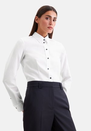 Femme portant une chemise blanche cintrée à boutons noirs et un pantalon noir taille haute sur un fond clair uni.