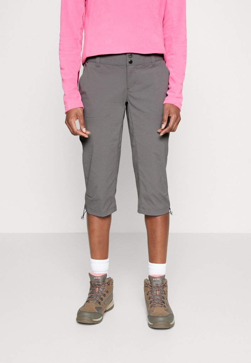 Pantalones capri grises con botones al frente y puños con cremallera, combinados con botas de montaña marrones y calcetines blancos. La parte superior es de color rosa y de mangas largas.