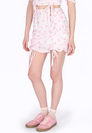 Frau trägt einen weißen Minirock mit pinkem Blumenmuster und seitlichen Bändern, kombiniert mit transparenten Knöchelsocken und pinken Sneakers.