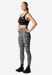Svart sports-BH og høyt liv leggings med et grått kamuflasjemønster. Modellen har på seg svarte joggesko med merkevaren sitt logo på siden.