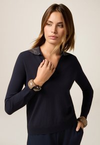 Jonge vrouw in een donkerblauwe trui met een glinsterende kraag, twee dikke armbanden, en kijkt lichtjes opzij met een hand bij haar nek.