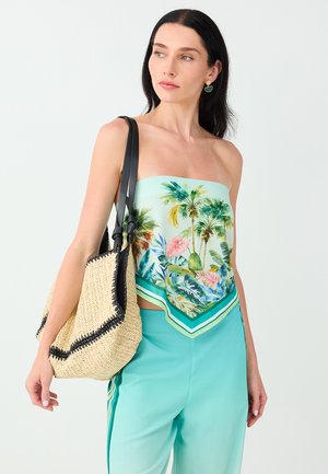 Vrouw draagt een strapless top met tropische print, turquoise broek, draagt een grote beige gevlochten tas met zwarte leren bandjes over haar schouder.