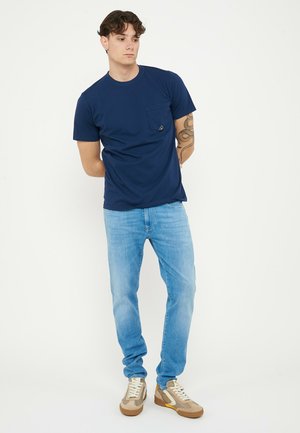 Giovane uomo con braccio tatuato che indossa una maglietta blu navy, jeans azzurri e sneakers beige, in piedi con le mani dietro la schiena.