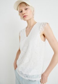 Hvit blondetanktopp med V-hals, med detaljerte blondedetaljer på skuldrene og et teksturert blomstermønster gjennom hele.