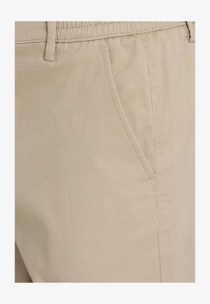 Beige katoen broek met een gestructureerde afwerking, voorzien van een zijzak en een elastische tailleband voor comfort. Close-up van de stofdetails.