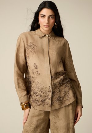 Donna con i capelli lunghi e scuri che indossa una camicia abbottonata color sabbia con motivo floreale e pantaloni coordinati, posando con una mano in tasca e un braccialetto al polso.