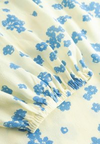 Tissu jaune clair avec motif floral bleu ; présente un élastique froncé au niveau de l'encolure, ajoutant texture et dimension.