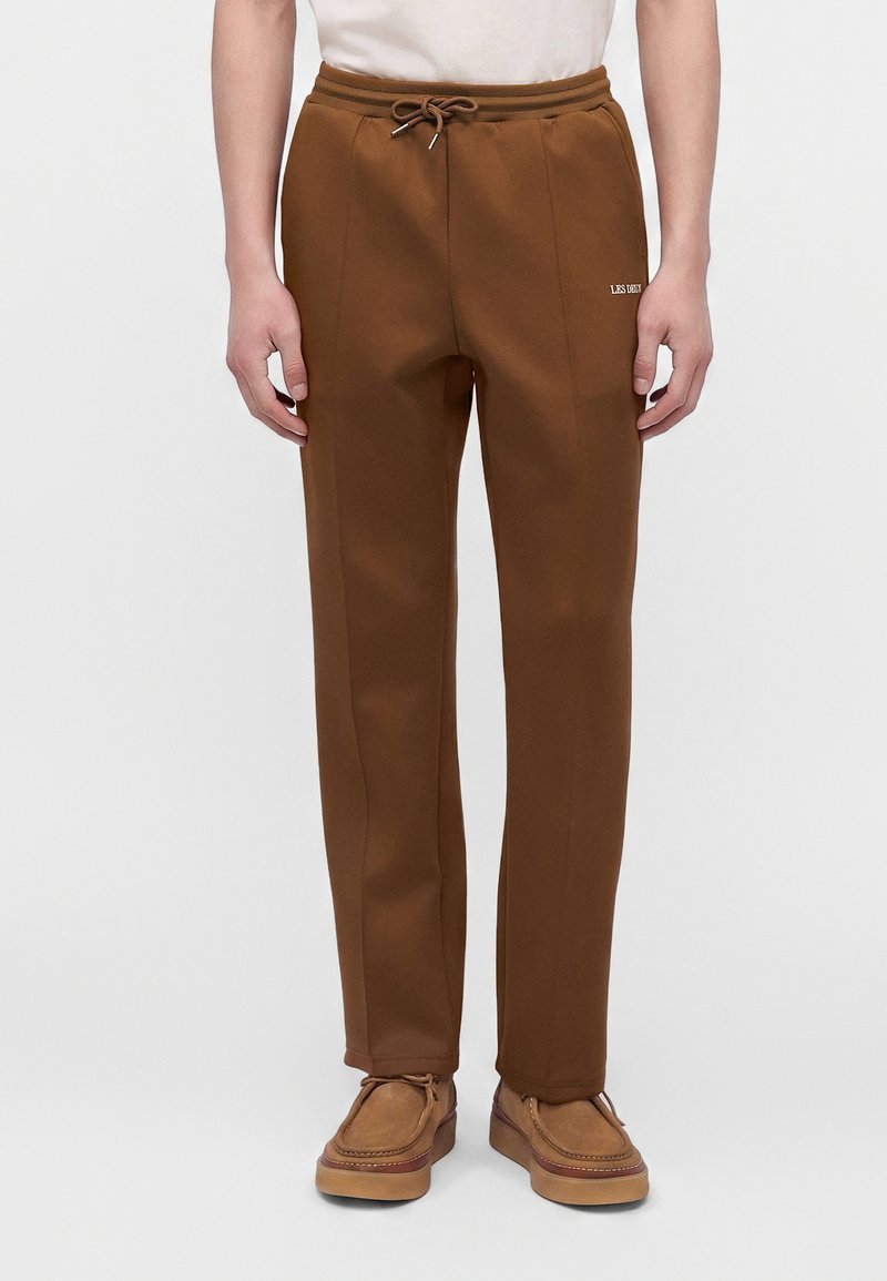Άνδρας που φοράει καφέ παντελόνι με ίσια πόδια και κορδόνι στη μέση, καθώς και καφέ δερμάτινα παπούτσια με κορδόνια, στέκεται μπροστά σε απλό φόντο.