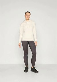 Hoodie beige chiaro con cordini, tasca frontale, abbinato a leggings grigi scuri e scarpe da ginnastica nere su uno sfondo neutro.