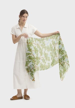 Robe en lin blanc avec un devant boutonné, associée à des sandales marron à enfiler. Le mannequin tient une écharpe transparente ornée de motifs d'arbres verts sur un fond clair.