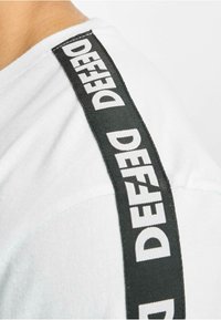 White T-shirt with black fabric shoulder tape featuring the word "DEFEAT" in bold white letters, smooth texture, and relaxed fit.