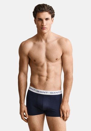 Junger Mann mit durchtrainiertem Körperbau, trägt dunkelblaue Boxershorts mit weißem Bund und der Marke "GANT" vor einem schlichten Hintergrund.