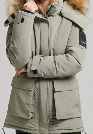 Personne portant une veste d’hiver matelassée vert clair avec une capuche bordée de fourrure, des poches à rabat et un patch noir sur le bras gauche.