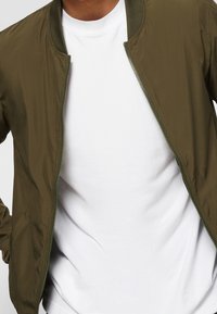 Oliwkowa bomberka z zamkiem z przodu, prążkowanym kołnierzem i gładkiego materiału, noszona na białym podkoszulku z okrągłym dekoltem.