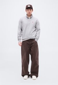 Νέος άνδρας που φορά καφέ καπέλο, ανοιχτό γκρι φούτερ με φερμουάρ, φαρδιά καφέ παντελόνια, λευκά αθλητικά παπούτσια, όρθιος μπροστά σε απλό λευκό φόντο.