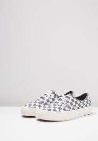 Ett par låga Vans-sneakers med svart och vitt rutmönster, vita snören och tjocka vita sulor på en enfärgad yta.