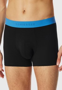Black cotton boxer shorts with a blue elastic waistband featuring the brand name "SCHIESSER" in a contrasting color. Smooth texture.