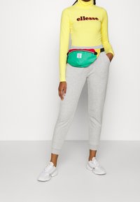 Grüne Bauchtasche mit gelben, roten und schwarzen Akzenten, getragen über einem gelben Rollkragenpullover und grauen Sweatpants, kombiniert mit weißen Turnschuhen.