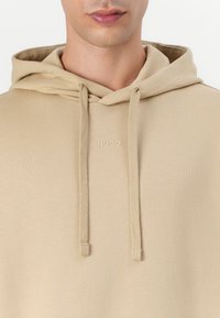 Sweatshirt bege com cordões, textura suave e logótipo "HUGO" em relevo no peito. Apresenta um design clássico de bolso canguru.