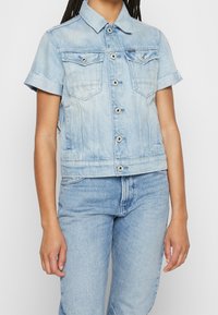 Ljusblå denimjacka med korta ärmar, två bröstfickor och silverdetaljer. Har en kortad design och slitna ärmslut.