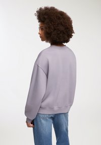 Przeskalowany fioletowy sweter z okrągłym dekoltem i opadającymi ramionami, w połączeniu z jasnoniebieskimi dżinsami. Materiał wydaje się miękki i teksturowany.