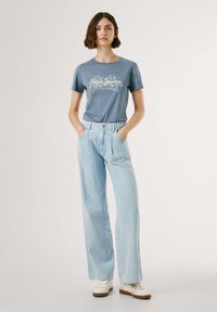 Gaiši zils t-krekls ar ziedu grafisko dizainu; apvienots ar gaiši ziliem augstās jostas beigām džinsiem; modelis valkā baltas sporta kurpes.